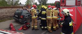 Strażacy i ratownicy medyczni transportują na noszach osobę poszkodowaną. W tle zniszczony samochód.