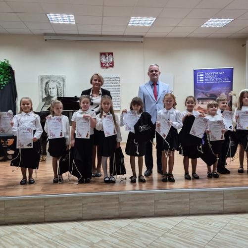 Uczniowie klas pierwszych cyklu sześcioletniego Państwowej Szkoły Muzycznej I stopnia im. Jana Sebastiana Bacha w Grajewie stoją na scenie z dyplomami, w towarzystwie nauczycieli. Dzieci ubrane są w stroje galowe(białe koszule i czarne spódnice/spodnie). W tle widoczne są fortepian, godło Polski oraz baner szkoły