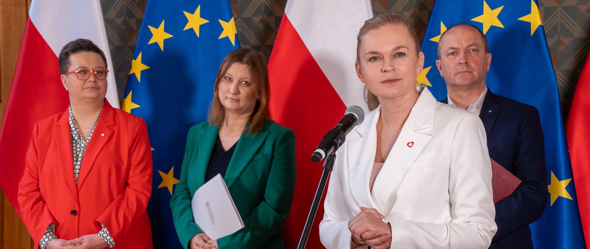 Przy ścianie na tle flag Polski i Unii Europejskiej stoją wiceministrowie, od lewej: wiceminister Katarzyna Lubnauer, Paulina Piechna-Więckiewicz, Henryk Kiepura. Przed nimi przy mikrofonie stoi minister Barbara Nowacka.
