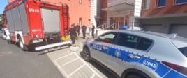 Na pierwszym planie widać pojazdy policji oraz straży pożarnej. Zdjęcie wykonane na zewnątrz, słoneczna pogoda. Widać stojących strażaków oraz policjantów. W tle budynki KPP w Wolsztynie oraz ulica.