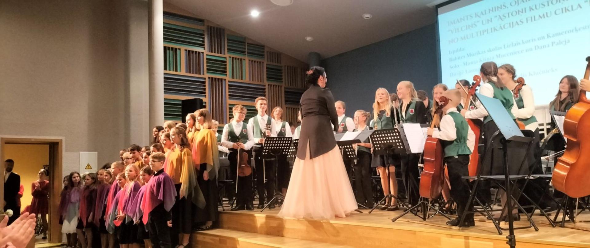 Cała scena zapełniona dziećmi i młodzieżą szkolną. Są chórzyści w pelerynkach, wielu muzyków z instrumentami smyczkowymi. Są też dwie dziewczyny, pewnie będą śpiewały. Przed nimi dyrygentka całego zespołu wokalno-instrumentalnego w czarnym fraku i długiej różowej sukni.
