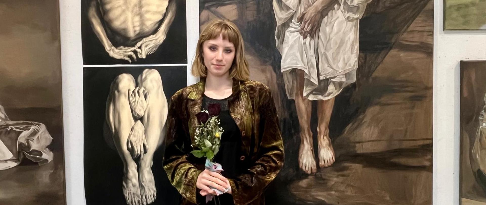 Na tle wiszących na szarym tle prac malarskich widać dziewczynę, ujętą w popiersiu z bukiecikiem kwiatków 