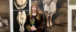 Na tle wiszących na szarym tle prac malarskich widać dziewczynę, ujętą w popiersiu z bukiecikiem kwiatków 