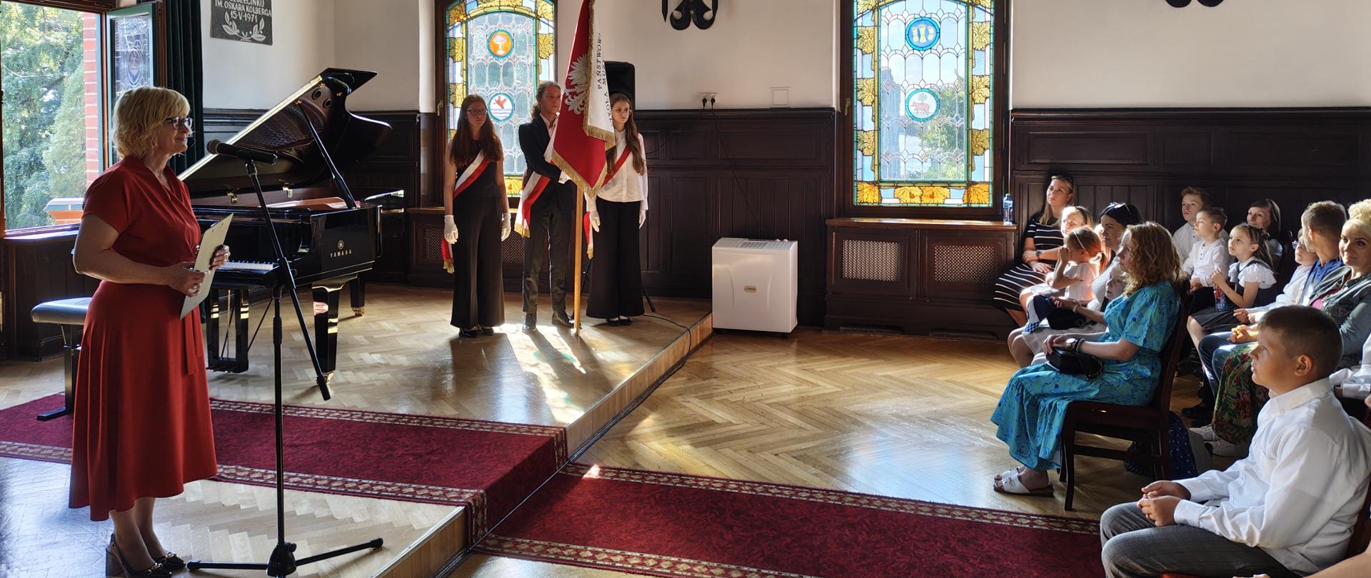 Kobieta w czerwonej sukience przemawia przy mikrofonie obok fortepianu, za nią stoi poczet sztandarowy, a po prawej stronie siedzi publiczność.