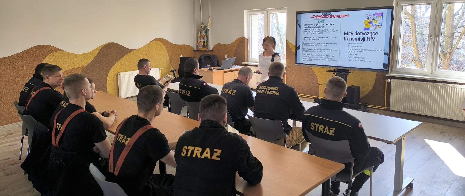 grupa funkcjonariuszy straży pożarnej siedzących w ławkach na szkoleniu i realizatorka z Inspekcji sanitarnej prowadząca zajęcia