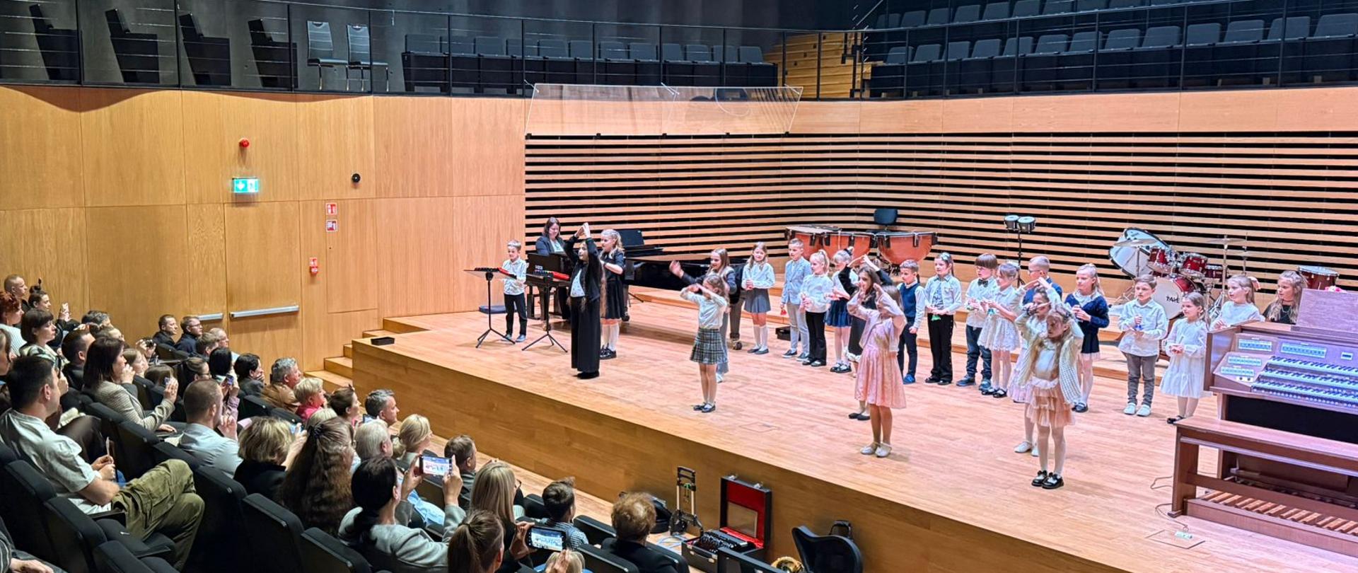 Dzieci ustawione w rzędach śpiewają i pokazują rękami gesty, kobieta akompaniuje na fortepianie. Z przodu widoczna publiczność.