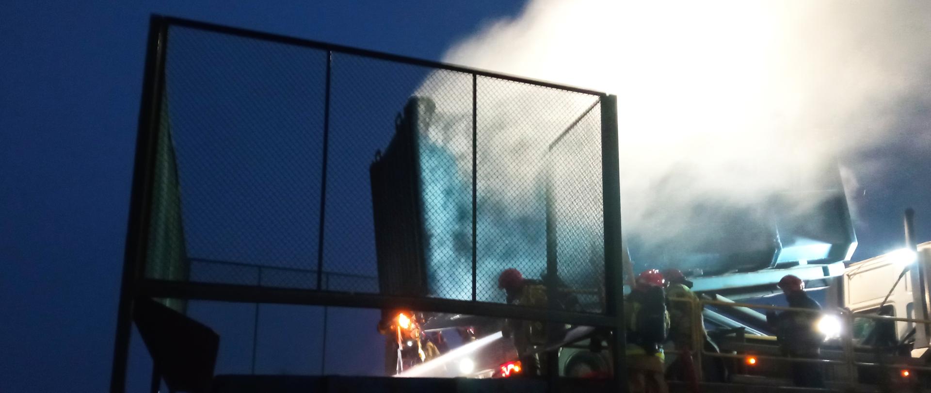 Zdjęcie przedstawia bezpieczne przesypywanie odpadów do innego kontenera (przy użyciu nośnika kontenerowego obsługiwanego przez pracownika stacji) z jednoczesnymi działaniami gaśniczymi prowadzącymi do likwidacji pożaru przez strażaków Komendy Powiatowej Państwowej Straży Pożarnej w Gołdapi. Na obrazie widocznych jest siedmiu funkcjonariuszy