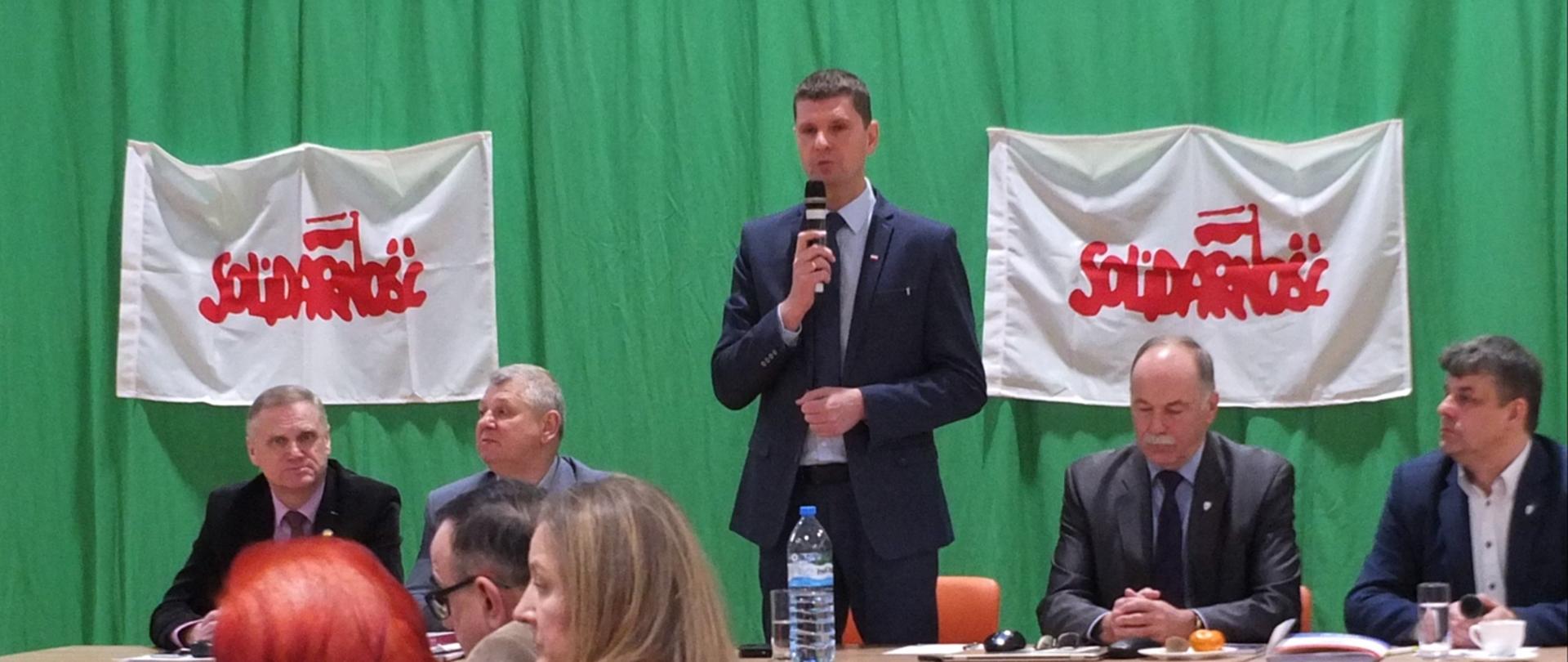 Czworo mężczyzn siedzących przy połączonych ławkach. Między nimi stoi z mikrofonem Minister Edukacji Narodowej – Dariusz Piontkowski. W tle zielony materiał i dwa transparenty z logo "Solidarność".