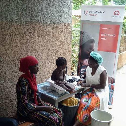 Czarnoskóra pielęgniarka waży czarnoskóre dziecko, obok którego siedzi jego mama. 