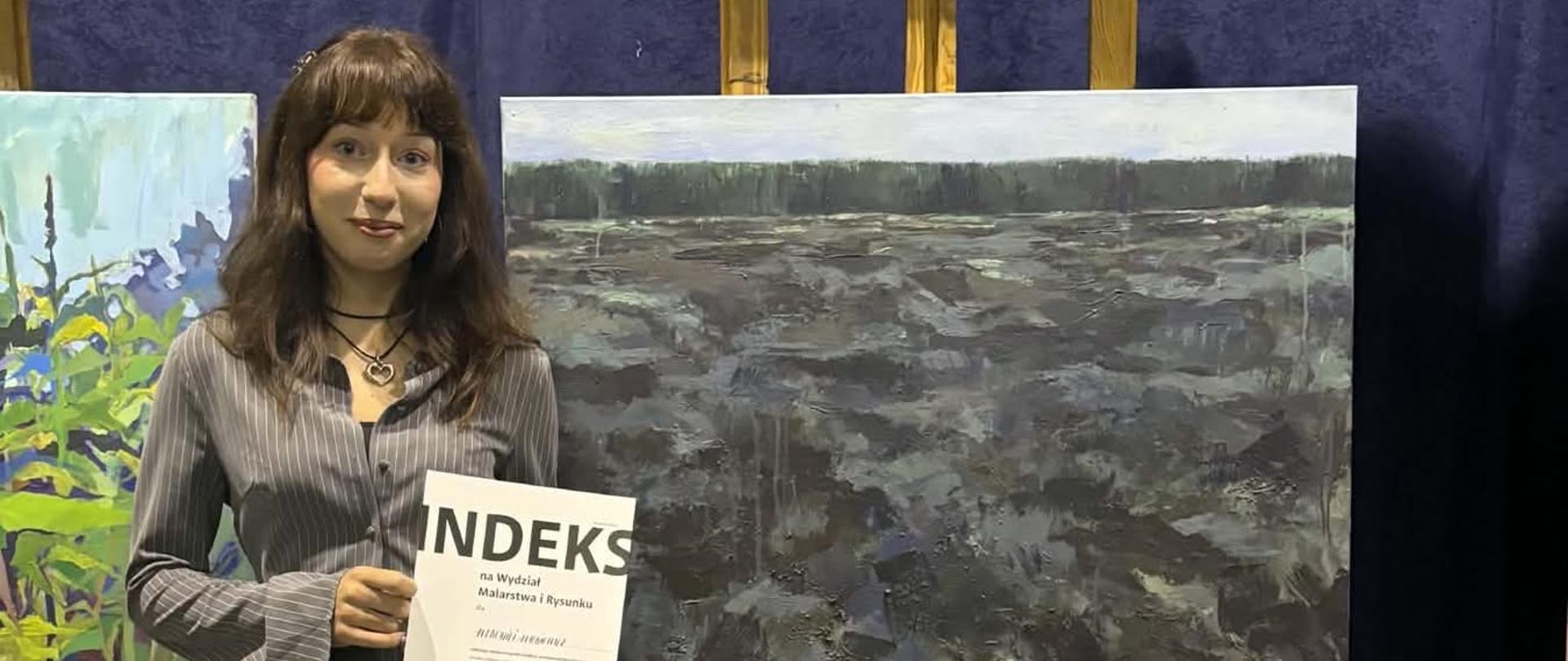 Młoda dziewczyna z dyplomem z dużym napisem INDEKS, za nią kilka obrazów na sztalugach