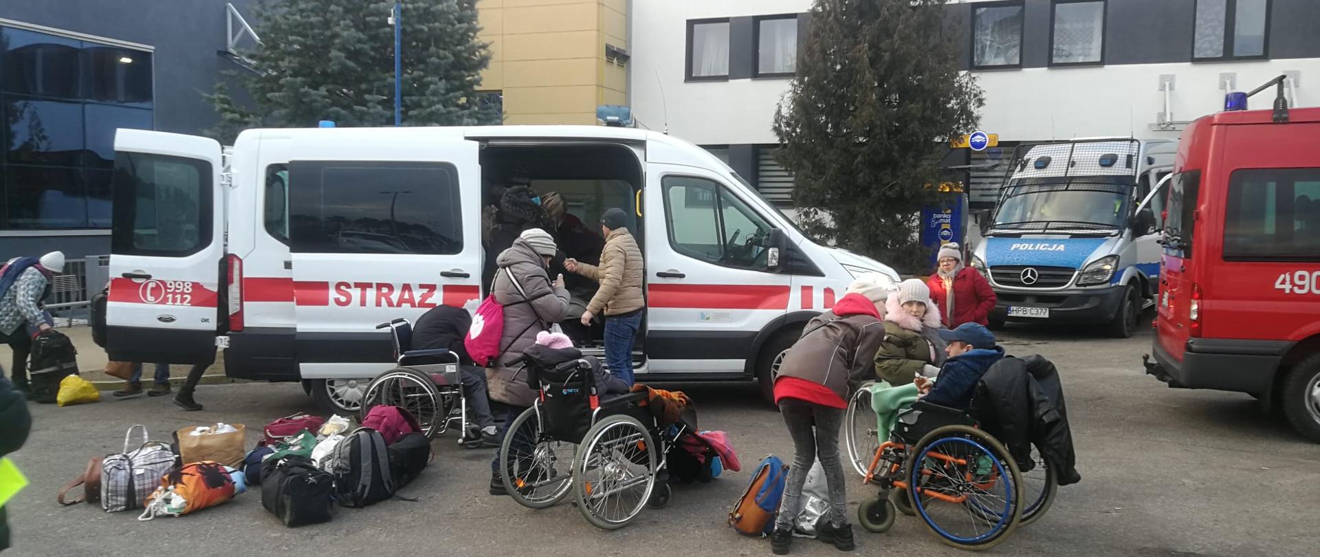 Zdjęcie przedstawia biały mikrobus Państwowej Straży Pożarnej do którego wsiadają uchodźcy z Ukrainy