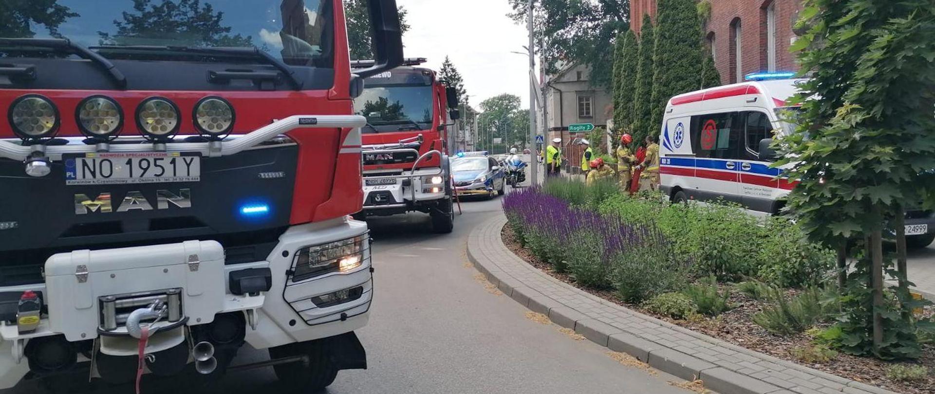Na zdjęciu z lewej stoją dwa samochody strażackie. Z prawej przy budynku karetka, za nią strażacy i ratownicy.