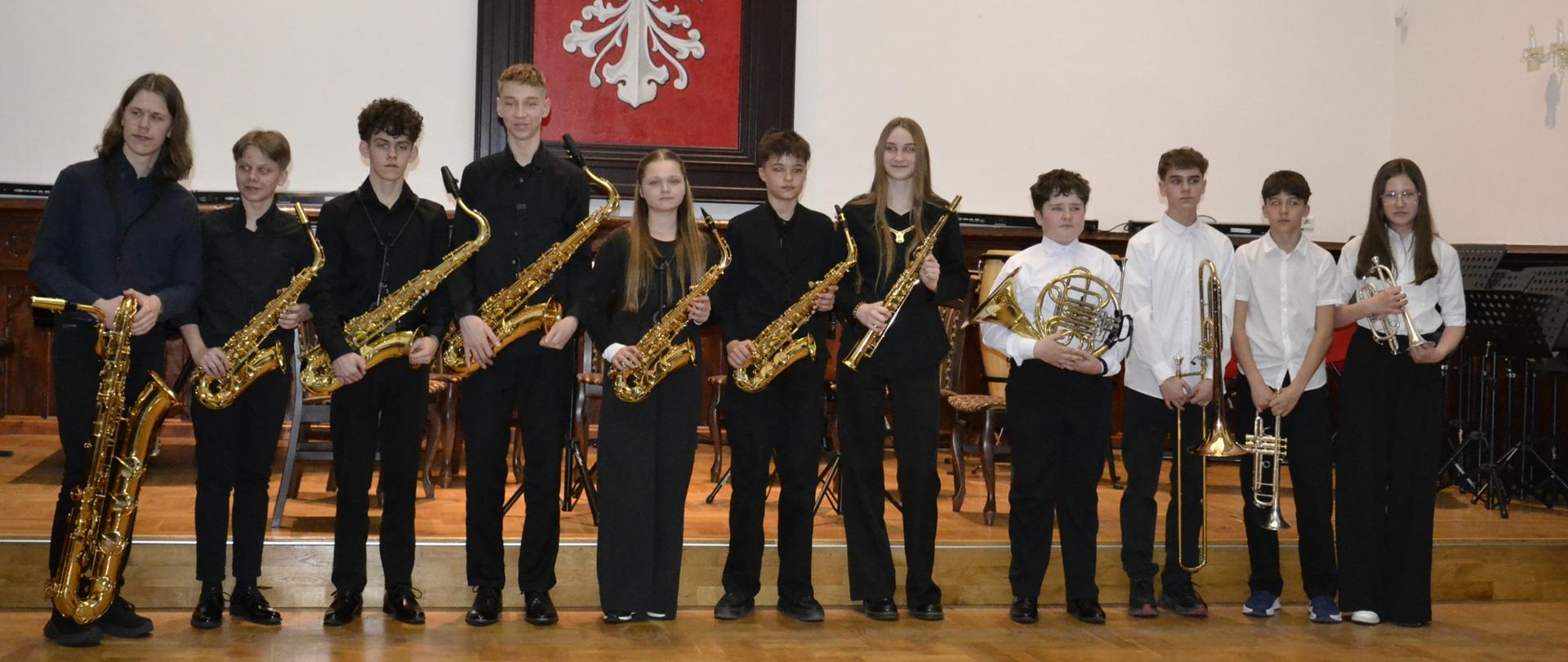 Grupa uczniów stojących na scenie w Sali Królewskiej z instrumentami dętymi – saksofonami, puzonem i trąbką – podczas koncertu zespołów kameralnych.