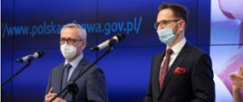 Ministrowie Marek Zagórski i Waldemar Buda podczas konferencji prasowej - stoją w maseczkach, przed mikrofonami.