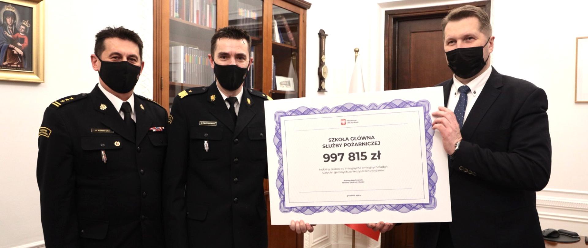 Minister Przemysław Czarnek przekazał SGSP czek na zakup specjalistycznej aparatury wykorzystywanej podczas pożarów, strażacy odbierają czek z rąk ministra Przemysława Czarnka.