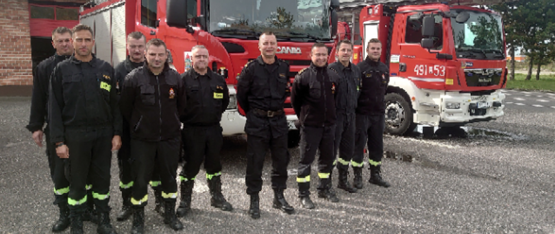 Na zdjęciu znajdują się strażacy w ubraniach koszarowych ustawieni na tle pojazdów pożarniczych i budynku komendy