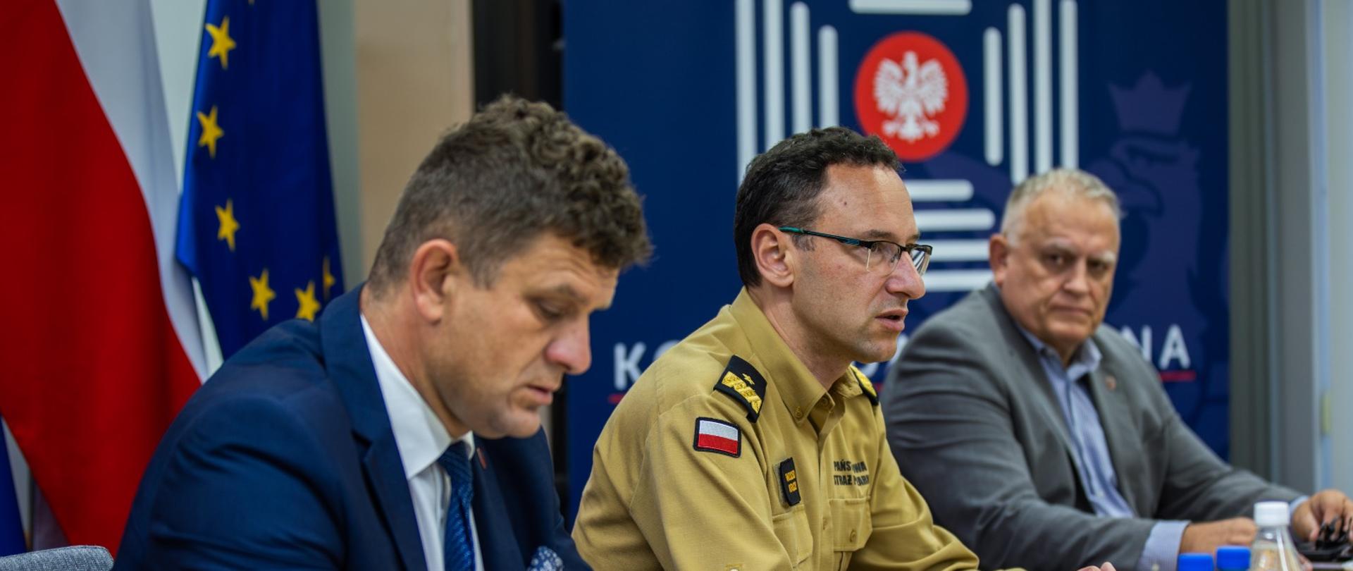 Trzech mężczyzn siedzi przy stole konferencyjnym, z czego jeden z nich – w żółtym służbowym mundurze PSP – mówi, gestykulując ręką. W tle widoczne są flagi Polski i Unii Europejskiej oraz baner z logotypem Komendy Głównej PSP.