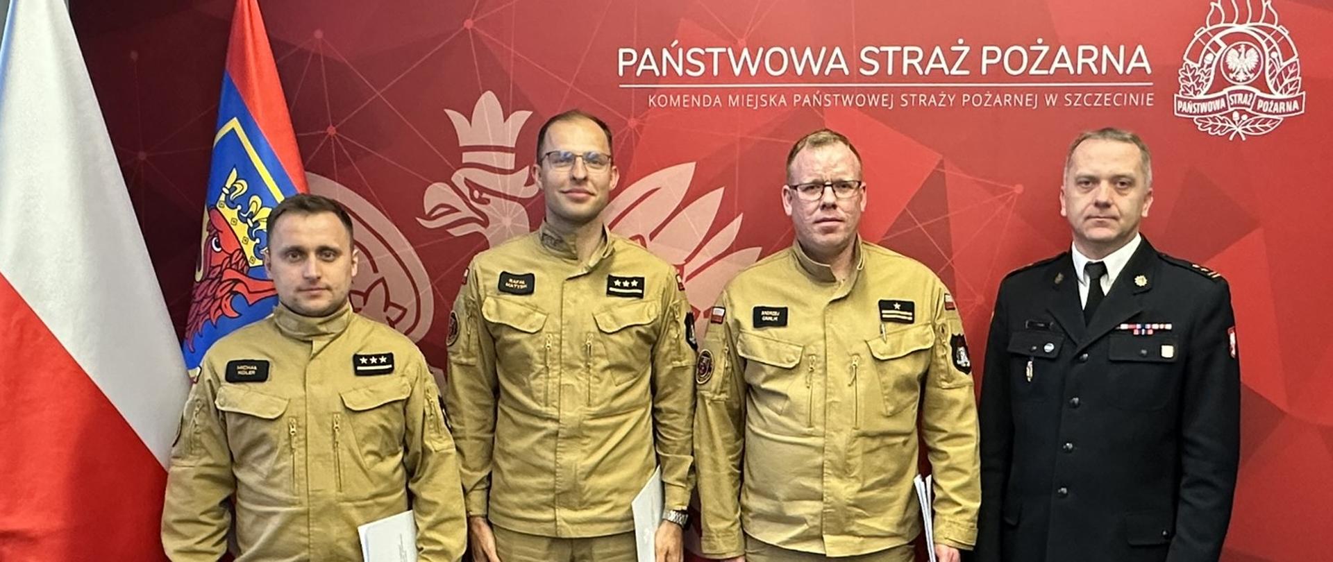 Czterech mężczyzn, trzech w mundurach bojowych straży pożarnej oraz jeden w mundurze wyjściowym, stoi na tle ścianki z herbem i napisem "Państwowa Straż Pożarna. Komenda Miejska Państwowej Straży Pożarnej w Szczecinie". Po lewej stronie widnieją flagi Polski oraz Województwa Zachodniopomorskiego