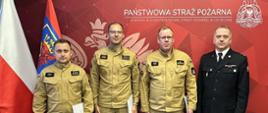 Czterech mężczyzn, trzech w mundurach bojowych straży pożarnej oraz jeden w mundurze wyjściowym, stoi na tle ścianki z herbem i napisem "Państwowa Straż Pożarna. Komenda Miejska Państwowej Straży Pożarnej w Szczecinie". Po lewej stronie widnieją flagi Polski oraz Województwa Zachodniopomorskiego