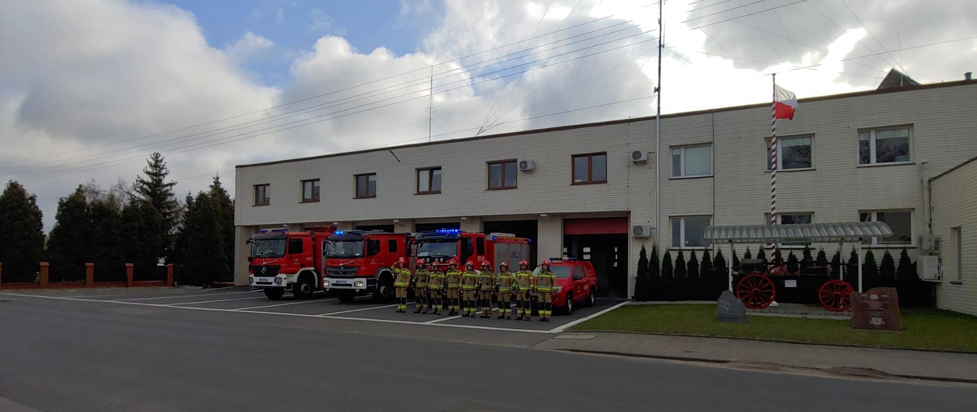 na zdjęciu jednostka ratowniczo gaśnicza cztery samochody pożarnicze wystawione są przed garaże, przed samochodami stoi dziewięciu strażaków na zbiórce