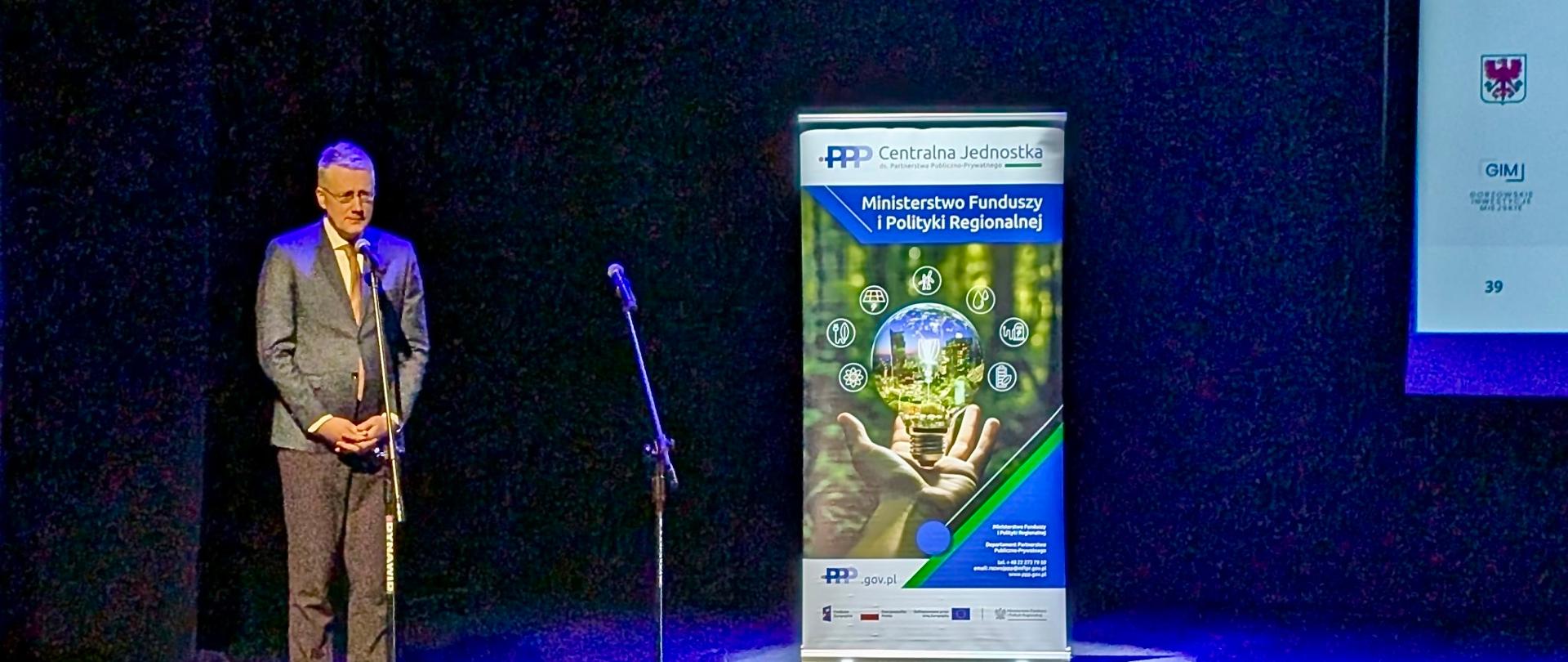 Na zdjęciu uczestnik konferencji stoi na scenie przed mikrofonem