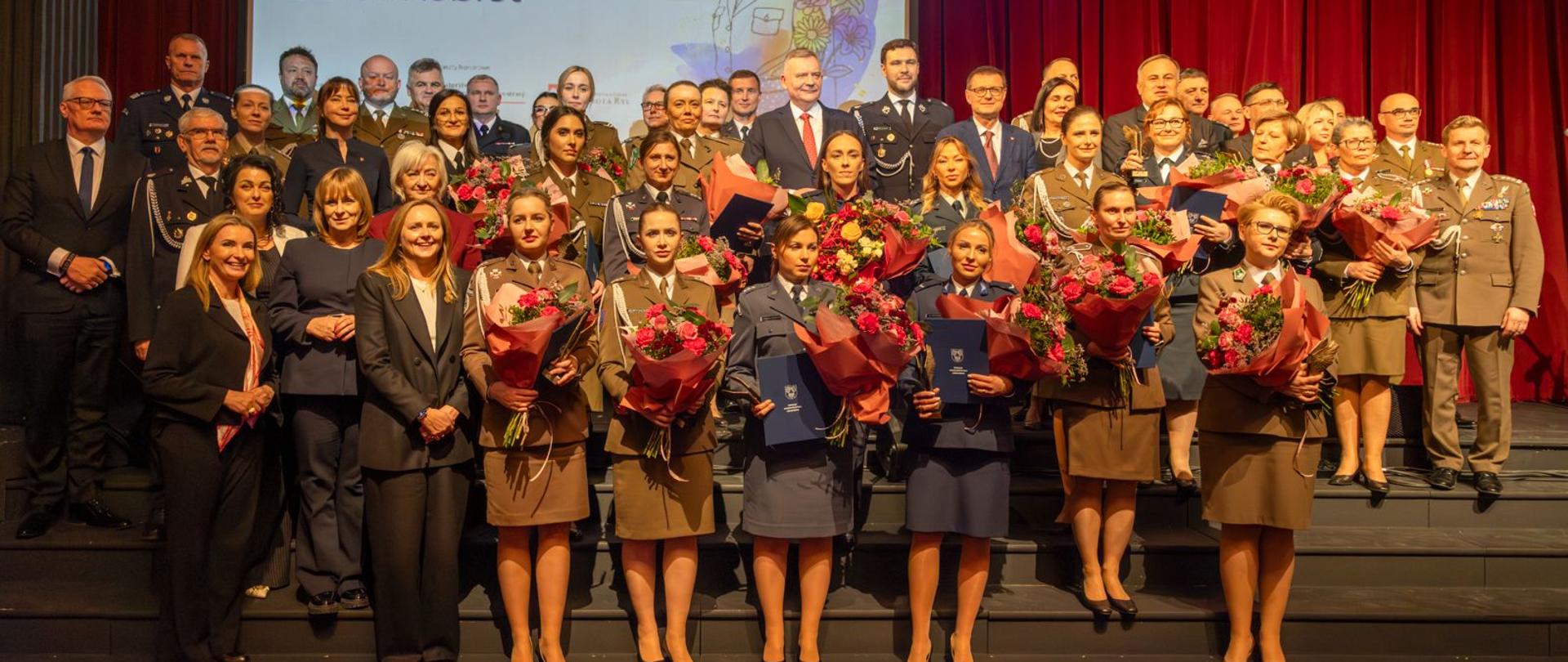 Uroczystości Mundurowego Dnia Kobiet funkcjonariuszki i zaproszeni goście wspólnie pozują do zdjęcia, stoją w kilku szeregach wyróżnione funkcjonariuszki trzymają w ręku kwiaty i listy pochwalne, okoliczności mają miejsce w Teatrze imienia Stefana Jaracza w Łodzi, za pozującymi widać ekran i wyświetlany na nim slajd z napisem Mudurowy Dzień Kobiet.