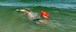Zdjęcie przedstawia osobę płynącą w morzu między falami, która ma przyczepiona do ciała unoszącą się na wodzie czerwona bojkę.
