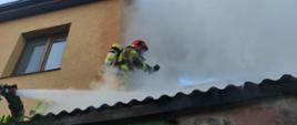 Zdjęcie przedstawia strażaka zabezpieczonego w sprzęt ochrony układu oddechowego, który stoi na dachu budynku. Na dachu widać duże zadymienie