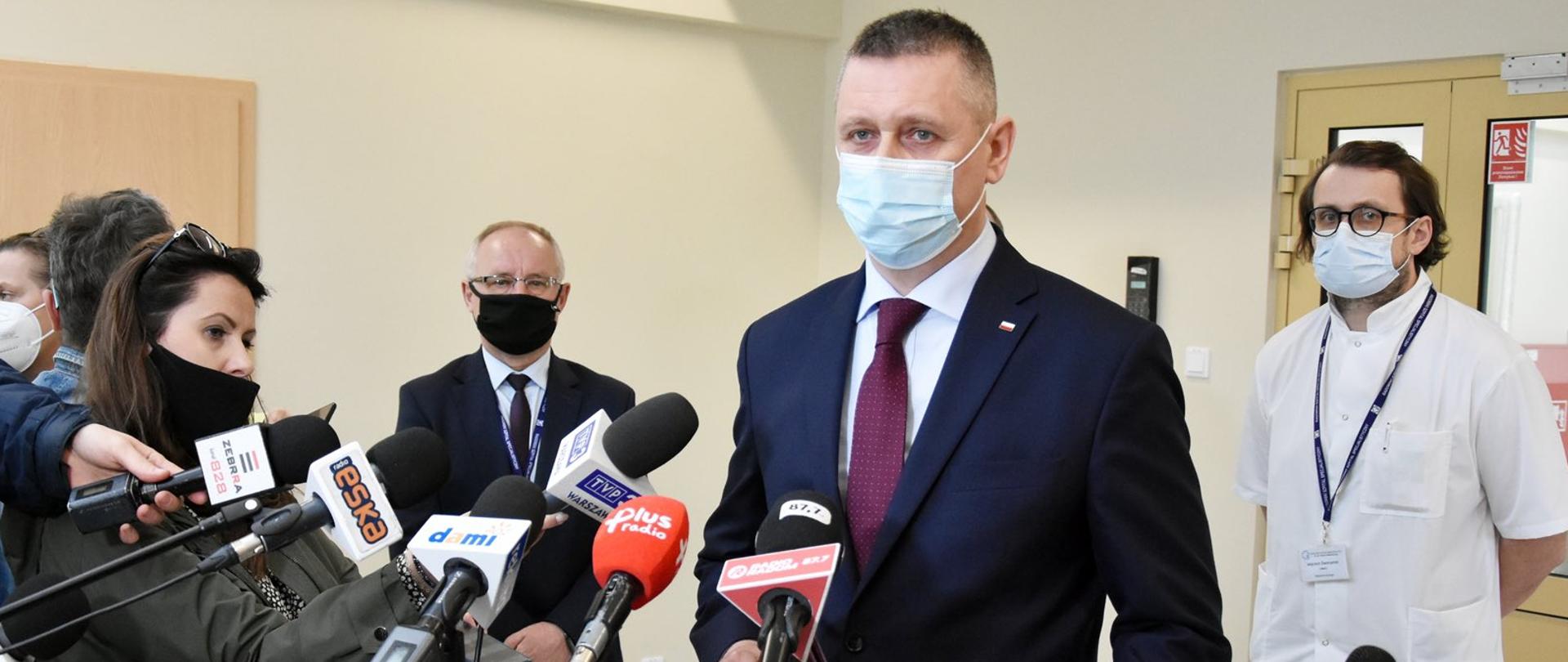 Elegancko ubrany mężczyzna przemawia podczas konferencji. Przed nim znajdują się mikrofony oraz dziennikarze. Na drugim planie znajduje się lekarz oraz drugi elegancko ubrany mężczyzna