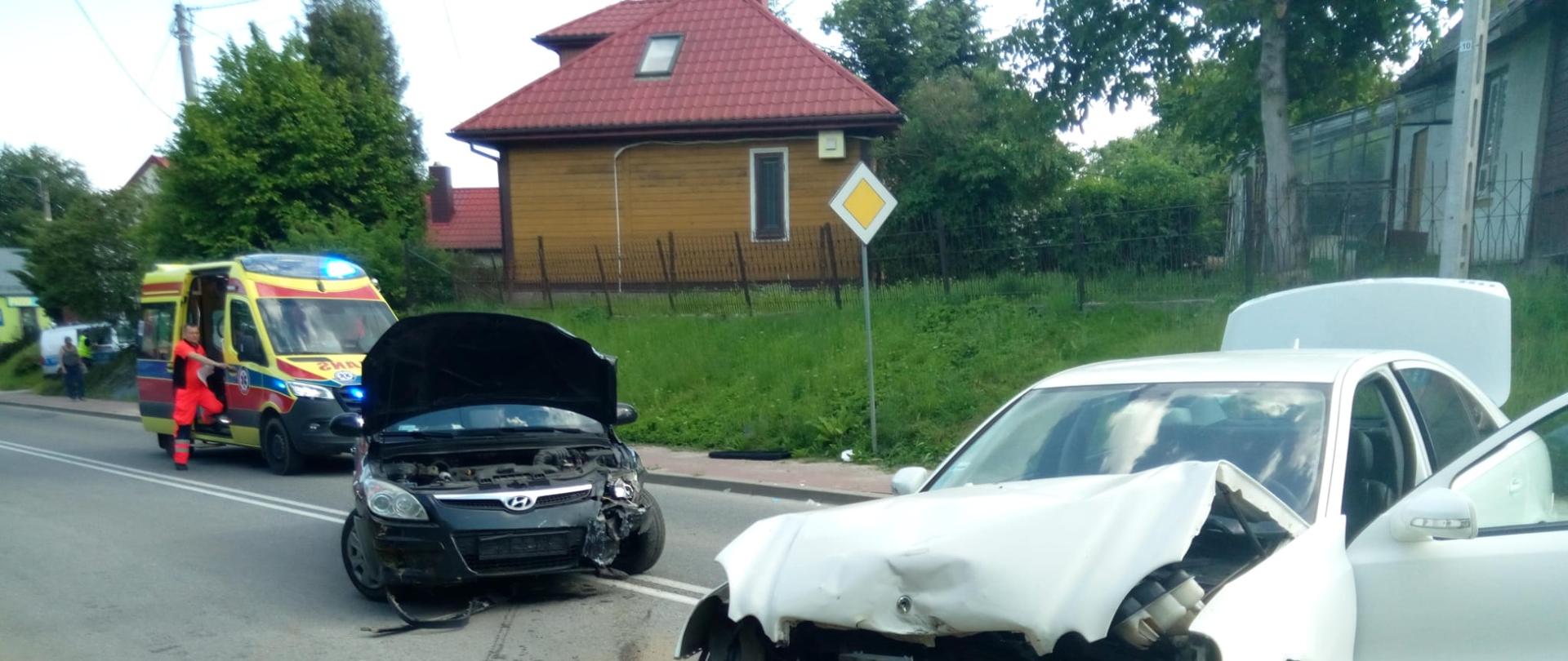 Zdjęcie przedstawia miejsce wypadku: dwa zniszczone samochody stoją na drodze, a w tle widać ambulans Zespołu Ratownictwa Medycznego.