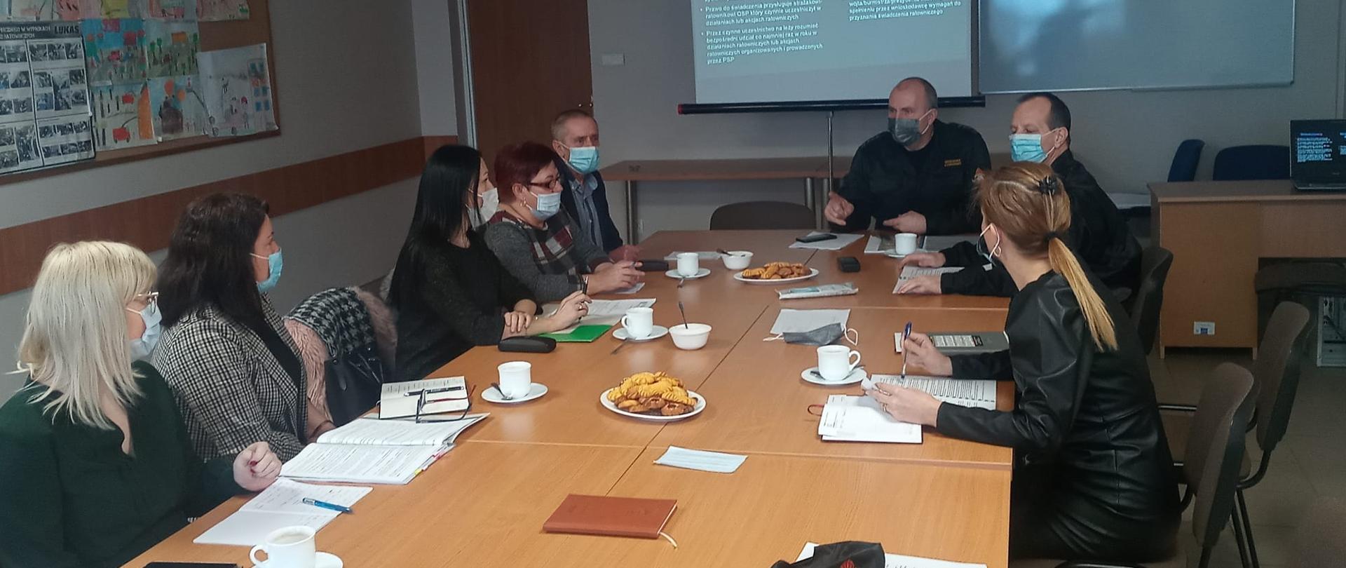 Zdjęcie przedstawia osiem osób siedzących przy brązowych stolikach, na których znajdują się filiżanki z kawą, dokumenty i dwa talerze z ciastkami. Znajduję się wewnątrz pomieszczenia. Na podłodze płytki w kolorze jasno brązowym. W tle na wprost znajduje się tablica, na której wyświetlony jest slajd z napisami. Po lewej stronie zdjęcia znajdują się brązowe drzwi. Nad nimi wisi zielona tabliczka oznaczająca wyjście ewakuacyjne.