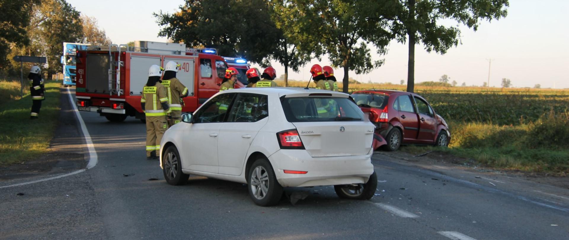 Uszkodozny smaochód marki Skoda z rozbitym tyłem blokuje jeden pas drogi,. Tuż za nim na poboczu citroen w kolorze czerwonym z uszkodzonym przodem oraz tyłem. Na drodze stoją strażacy realizujący działania. Droga została całkowicie zablokowana przez pojazdy pożarnicze.