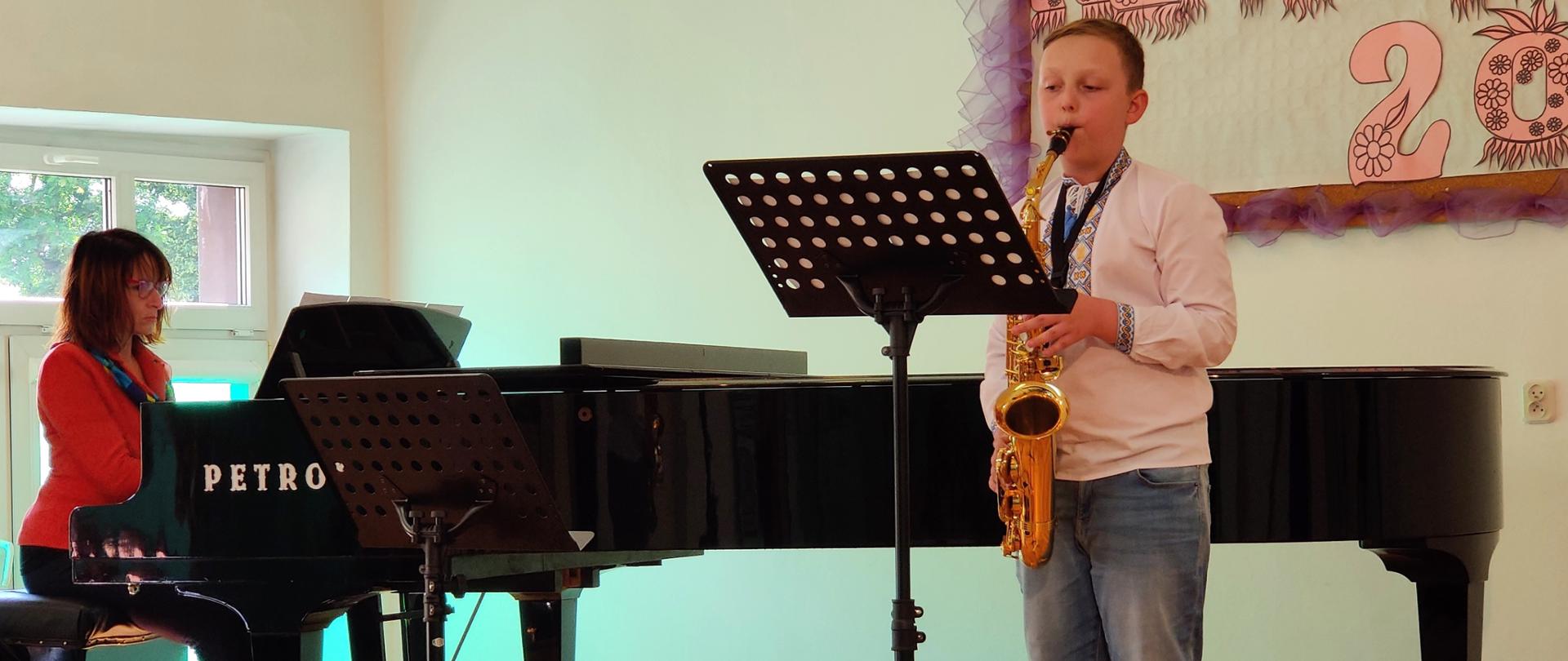 Zdjęcie przedstawiające jednego z wykonawców wraz z akompaniamentem fortepianowym pani Hałyny Hachak podczas koncertu uczniów klas pierwszych w dniu 14 czerwca 2023 r. na scenie w auli szkoły.