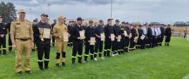 finałową ceremonię Gminnych Zawodów Sportowo-Pożarniczych jednostek OSP z terenu gminy Jabłonna Lacka 2026:
Uroczysta dekoracja: Na zdjęciu widoczny jest moment końcowy zawodów, w którym wyróżnieni strażacy ochotnicy ustawieni są w szeregu, trzymając w rękach otrzymane dyplomy oraz statuetki.
Uczestnicy: W szeregu znajdują się druhowie w różnorodnych mundurach – od piaskowych mundurów bojowych, przez czarne mundury koszarowe, aż po elementy umundurowania galowego.
Miejsce wydarzenia: Uroczystość odbywa się na płycie boiska sportowego, co podkreśla sportowy charakter rywalizacji.
Atmosfera: Kadr dokumentuje moment sukcesu i satysfakcji zawodników, którzy z dumą prezentują swoje osiągnięcia przed zgromadzonymi.
