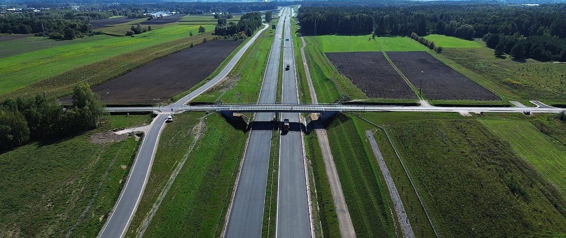 Widok z lotu ptaka na dwujezdniowy odcinek drogi ekspresowej S19. Nad trasą przebiega wiadukt, a po obu stronach znajdują się tereny zielone.
