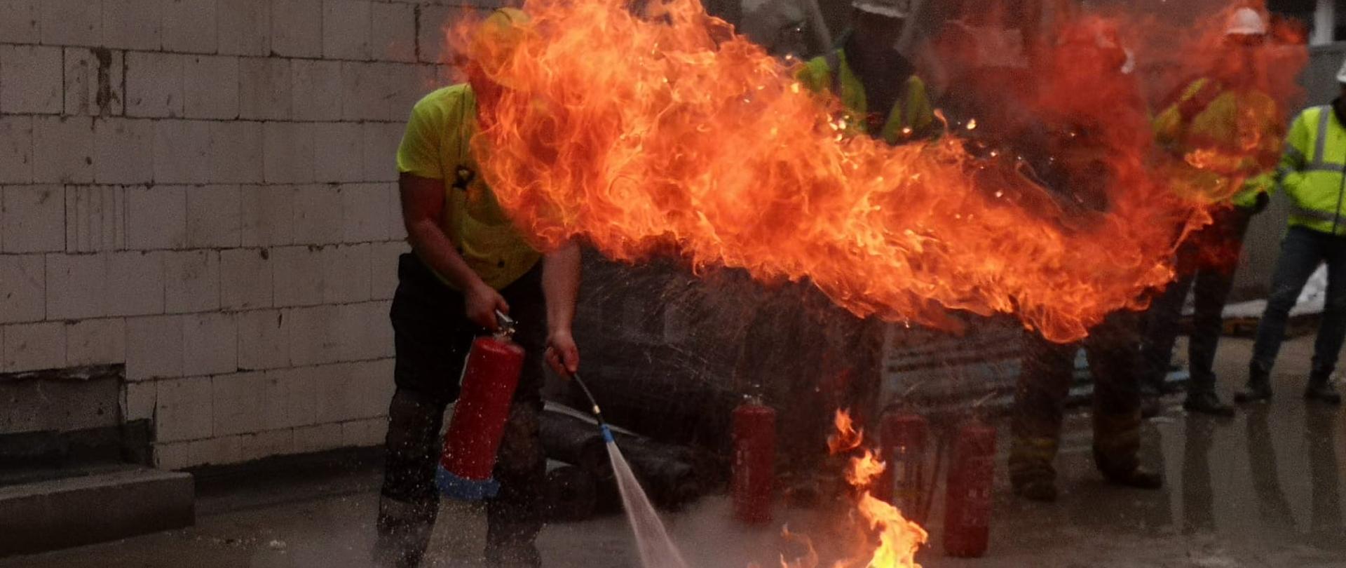 W środkowej części zdjęcia taca z rozpaloną cieczą stojąca na placu przy budowanym budynku, za tacą stoi jeden z pracowników z gaśnicą w ręku. W tle pozostałe osoby z budowy przyglądające się gaszeniu. 