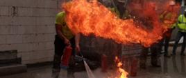 W środkowej części zdjęcia taca z rozpaloną cieczą stojąca na placu przy budowanym budynku, za tacą stoi jeden z pracowników z gaśnicą w ręku. W tle pozostałe osoby z budowy przyglądające się gaszeniu. 