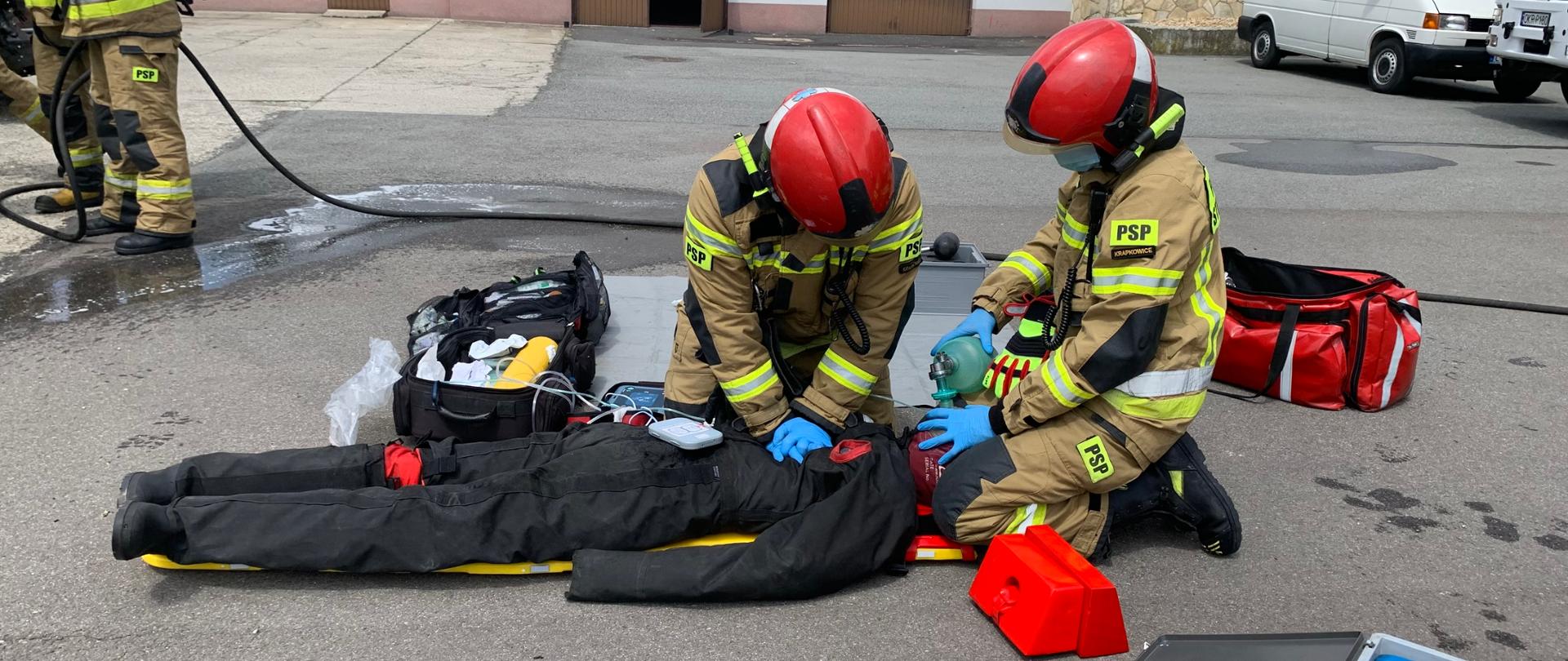 Na noszach ułożony manekin wydobyty spod samochodu. Dwóch ratowników PSP prowadzi resuscytację. Wokół rozłożony sprzęt medyczny.