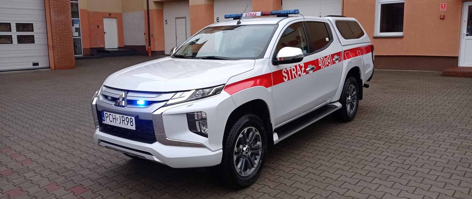Zdjęcie przedstawia nowy samochód strażacki osobowy specjalny Mitsubishi L200.
W tle budynek Komendy.
