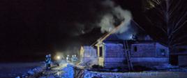 Wieczorem na zaśnieżonym podwórku stoją strażacy z rozwiniętymi wężami. Po prawej stronie budynek mieszkalny z którego wydobywa się dym.