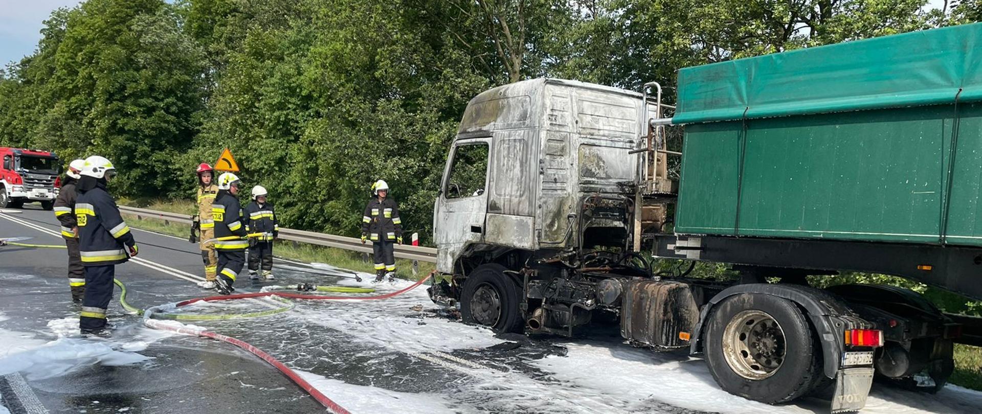 Na zdjęciu widać spalony ciągnik siodłowy z naczepą. Zdjęcie na drodze publicznej. WW ciągu dnia. Wokół pojazdu widać środki gaśnicze oraz linie gaśnicze.