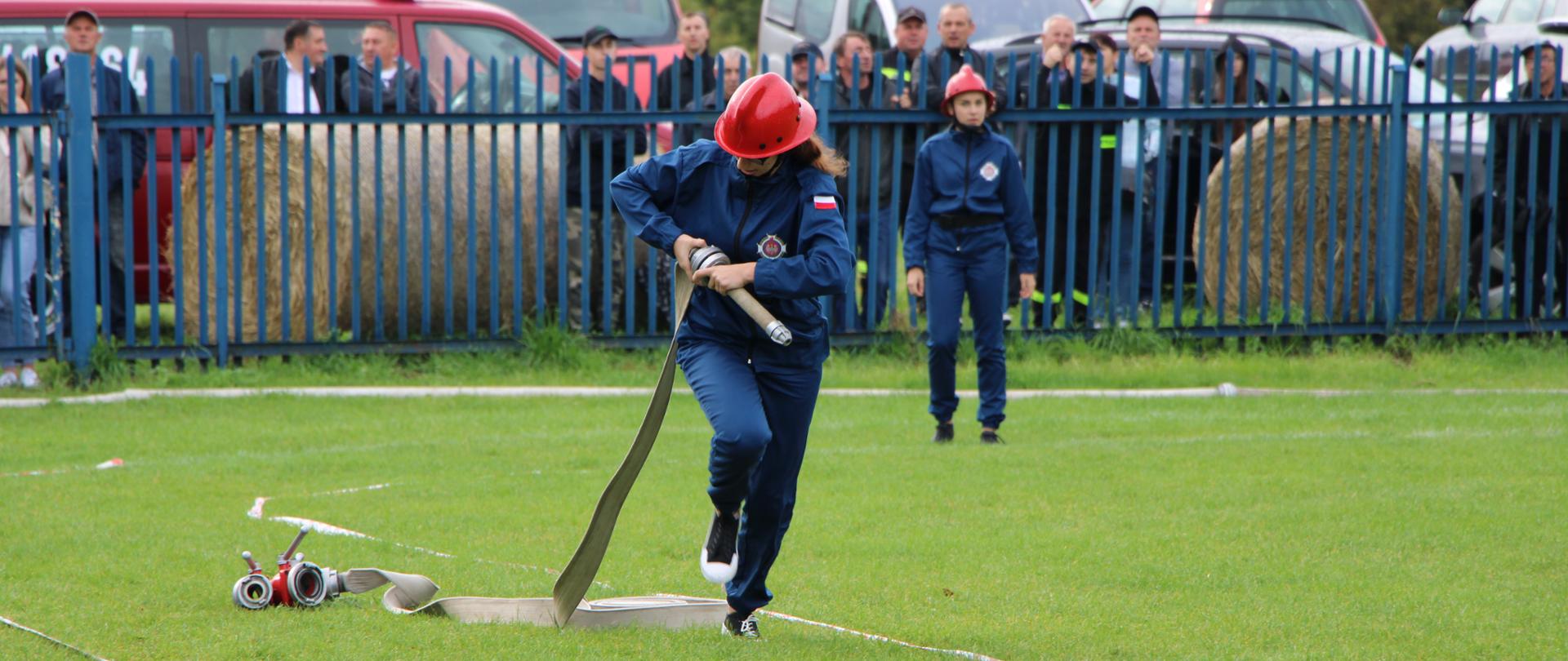 Zdjęcie obrazuje kobietę w stroju sportowym, która biegnąć podłącza prądownicę do węża. Zdjęcie zrobione podczas zawodów sportowo-pożarniczym. Na drugim planie, za metalowym ogrodzeniem znajdują się kibice, którzy bacznie obserwują poczynania zawodniczek.