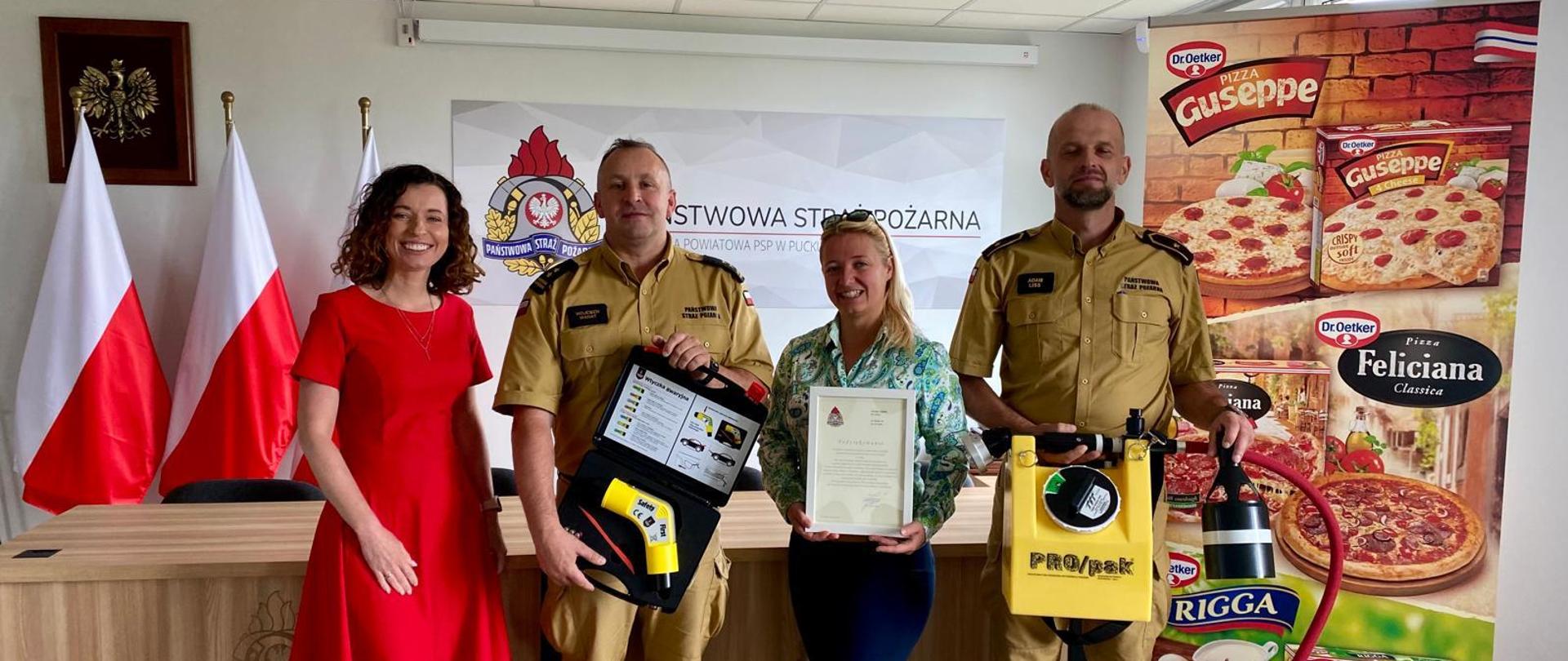 Zdjęcie wykonane w budynku Komendy Powiatowej PSP w Pucku. Na zdjęciu przedstawicielki Dyrekcji Zakładu Dr. Oetker Polska Sp. z o.o. oddział w Łebczu przekazują na ręce Komendanta Powiatowego PSP w Pucku Pana bryg. Wojciecha Wanata oraz Z-ca Dowódcy JRG w Pucku Pana asp. Adama Liss sprzęt ratowniczo gaśniczy. Z prawej strony reklama w postaci plakatu firmy Dr. Oetker. Na drugim planie z lewej strony flagi Rzeczpospolitej Polski oraz napis ,, Państwowa Straż Pożarna"