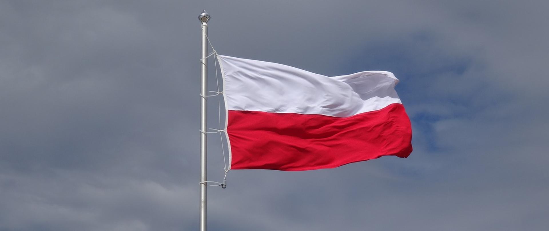 Zdjęcie przedstawia biało – czerwoną flagę Polski zawieszoną na białym maszcie powiewającą na wietrze. Tło zdjęcia stanowi niebo w kolorze niebieskim z białymi obłokami. 