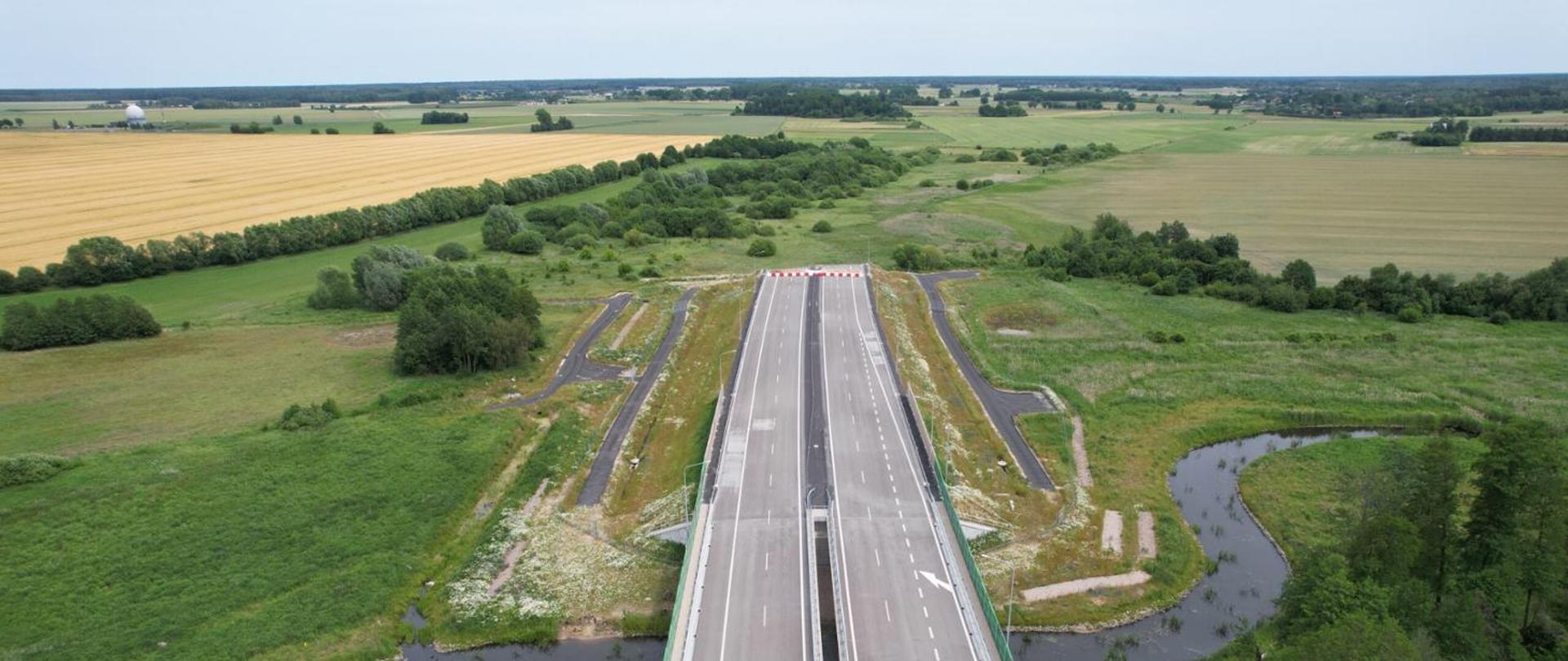 Widok z lotu ptaka na nowo wybudowany odcinek drogi z mostem nad małą rzeką, otoczony zielonymi polami i uprawami, w tle rozległy krajobraz wiejski.