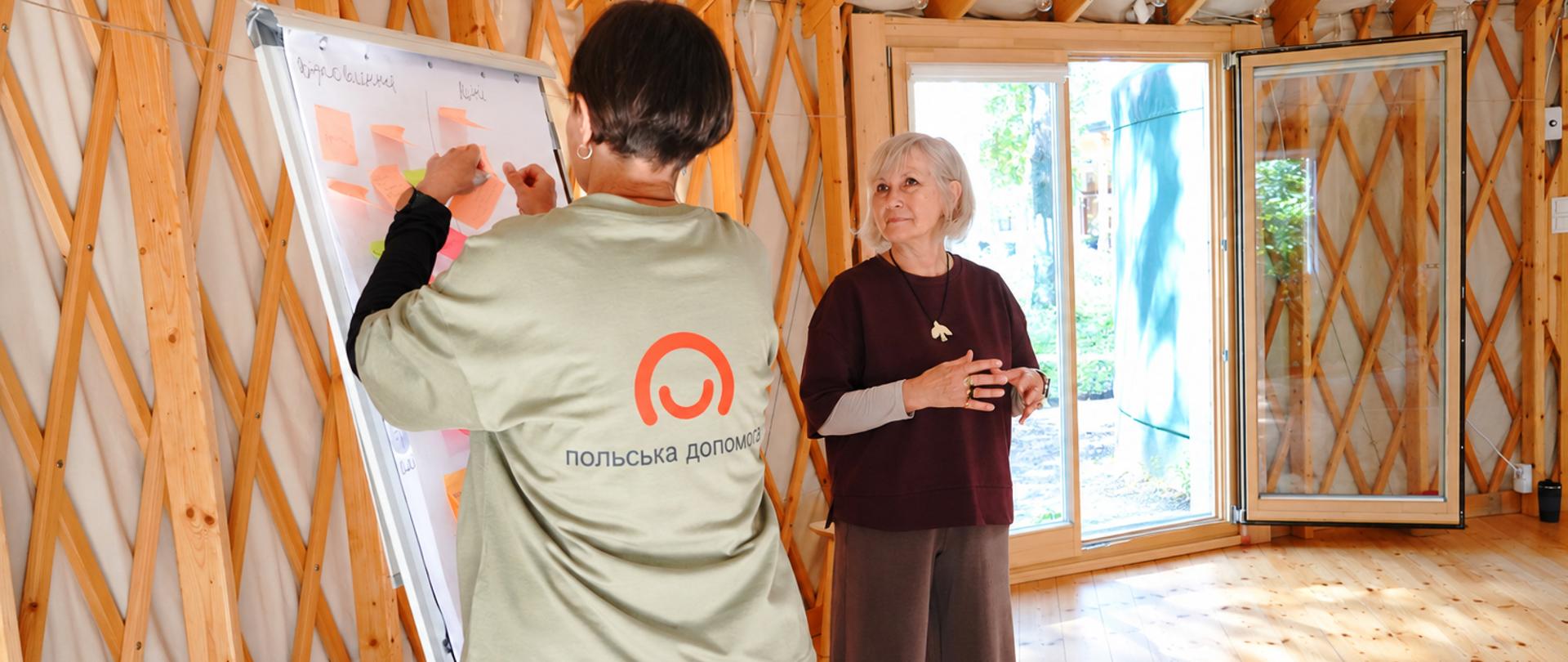 Dwie osoby pracują przy tablicy ustawionej w jasnej sali o drewnianej konstrukcji. Osoba stojąca tyłem przyczepia kolorowe karteczki do arkusza, a druga omawia wykonywane zadanie. Na koszulce osoby przy tablicy widoczne są znak programu Polska pomoc i napis w języku ukraińskim „Польська допомога”.