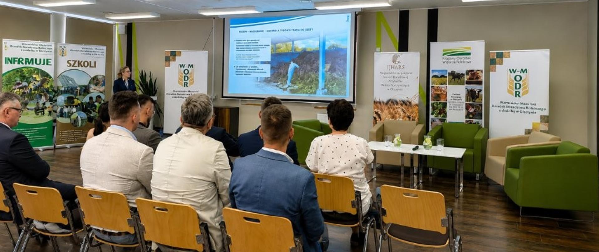 Kobieta stoi przy mównicy zwrócona do ekranu z prezentacją, przed nią grupa ludzi w rzędach krzeseł
