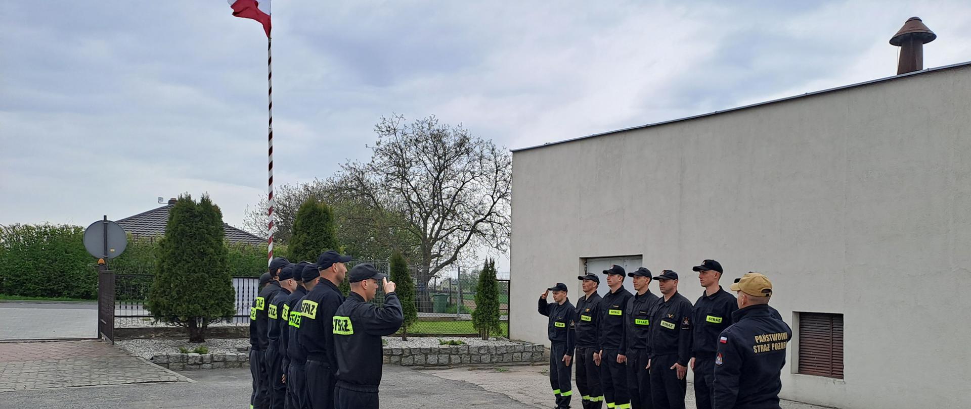 Zdjęcie przedstawia plac Jednostki Ratowniczo-Gaśniczej Komendy Powiatowej Państwowej Straży Pożarnej w Radziejowie, na którym odbywa się uroczysta zbiórka z podniesieniem Flagi Państwowej. Na zdjęciu strażacy ze zmiany przyjmującej oraz zdającej służbę.