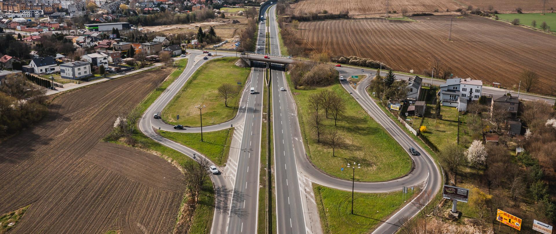 Zdjęcie przedstawia węzeł drogowy, który jest skrzyżowaniem drogi krajowej DK86 z ulicą Wolności w Będzinie. Na fotografii widoczna jest szeroka droga krajowa, która przechodzi przez most nad ulicą Wolności. Wokół skrzyżowania znajdują się pola uprawne oraz zabudowania mieszkalne. Na zdjęciu widać również kilka samochodów poruszających się po drogach. W tle można dostrzec zabudowania miejskie oraz przemysłowe. Fotografię wykonano z lotu ptaka, co pozwala na dokładne zobrazowanie układu dróg oraz otoczenia. W rym rejonie powstaną ekrany akustyczne.. 