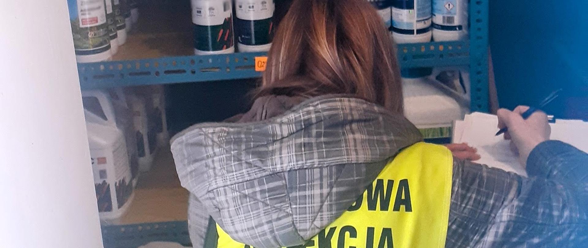 osoba w kamizelce odblaskowej z napisem "PAŃSTWOWA INSPEKCJA OCHRONY ROŚLIN I NASIENNICTWA" przeprowadza kontrolę śor na regale
